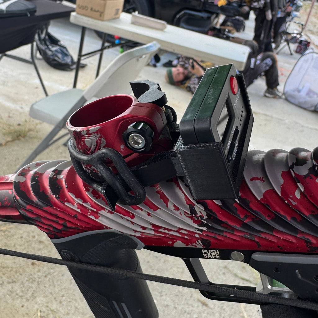 A close-up of a red and black paintball marker with a mounted black digital display, photographed outdoors at what appears to be an event or workshop.